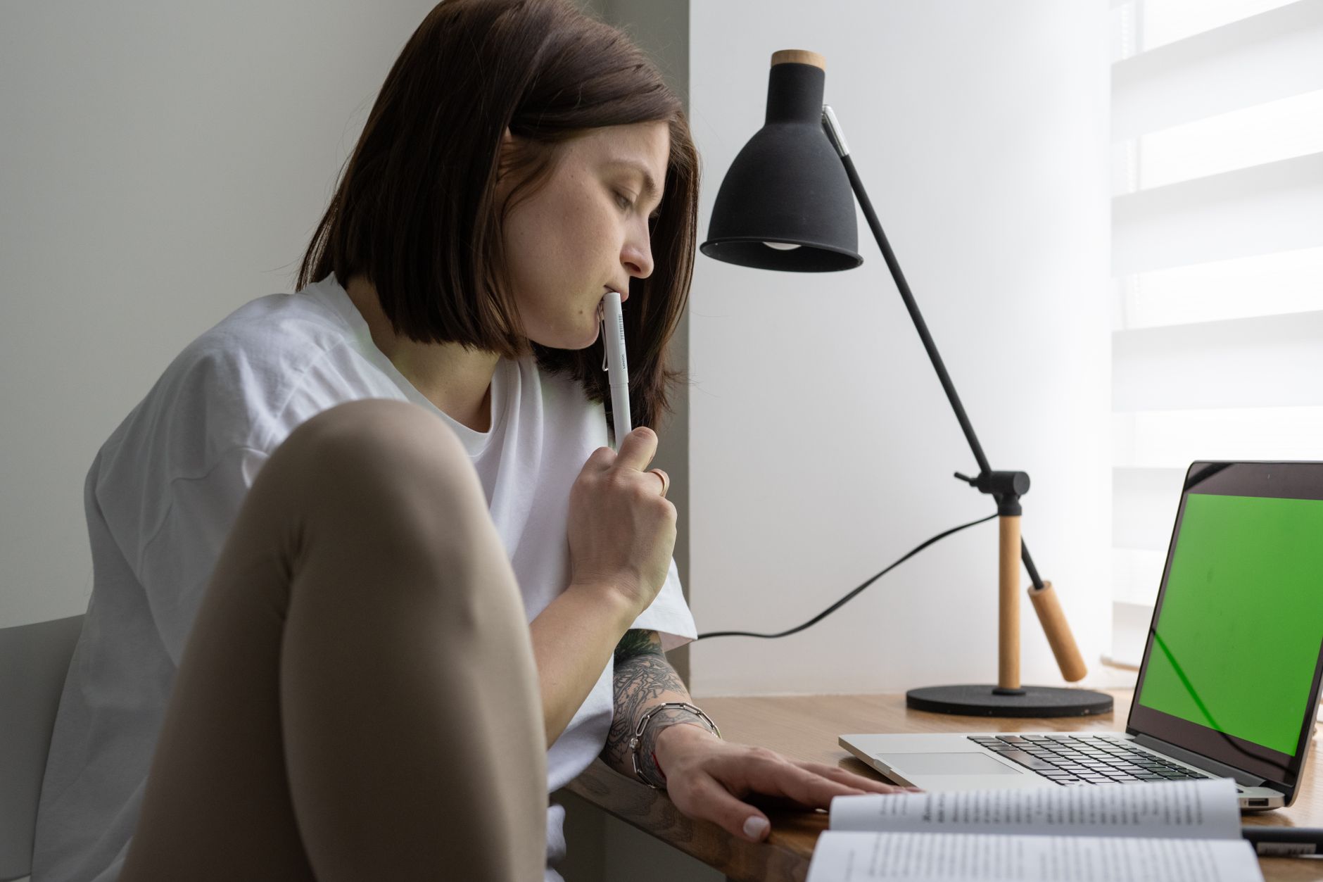 Side view of a thoughtful student with a pen, studying at a table with a notebook computer in a lit room.